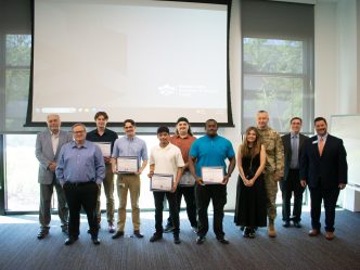 A group of men and one woman stand in front of a screen and hold awards.
