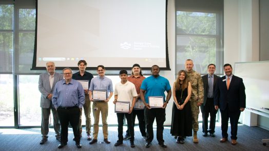 A group of men and one woman stand in front of a screen and hold awards.