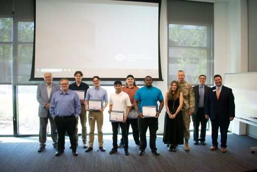A group of men and one woman stand in front of a screen and hold awards.