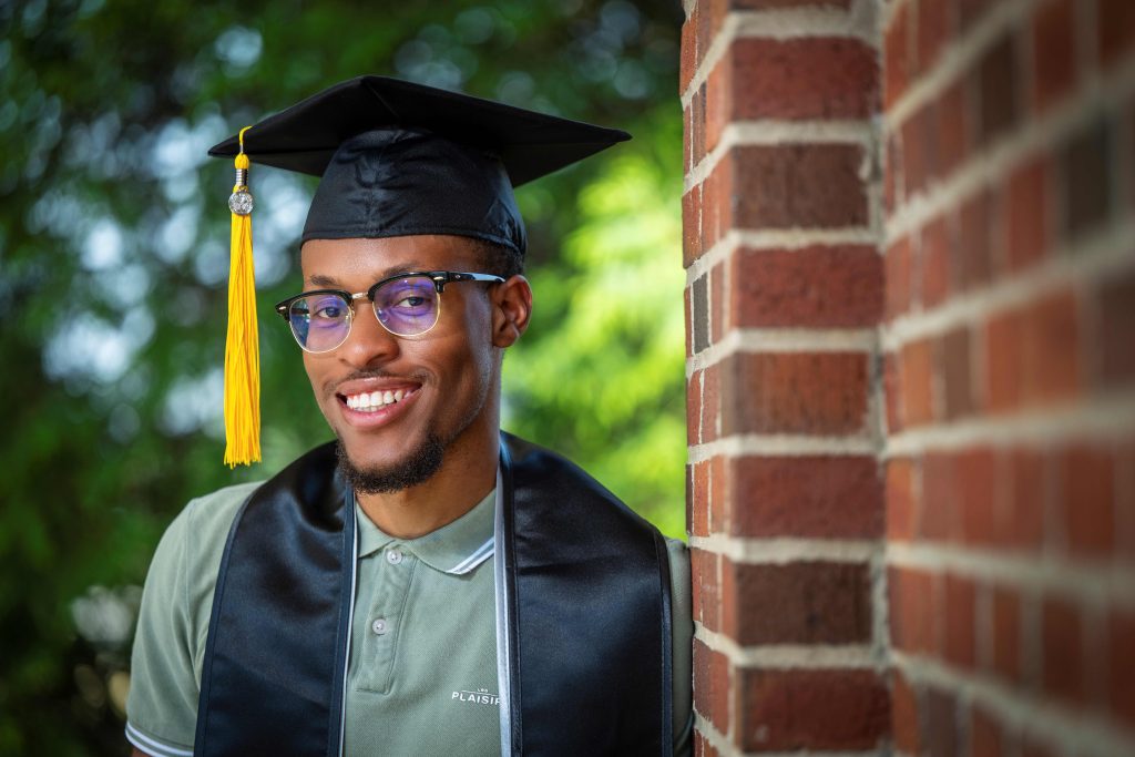 A man wearing a graduate hood and gown, while leaning against a brick wall, smiles for a photo. 
