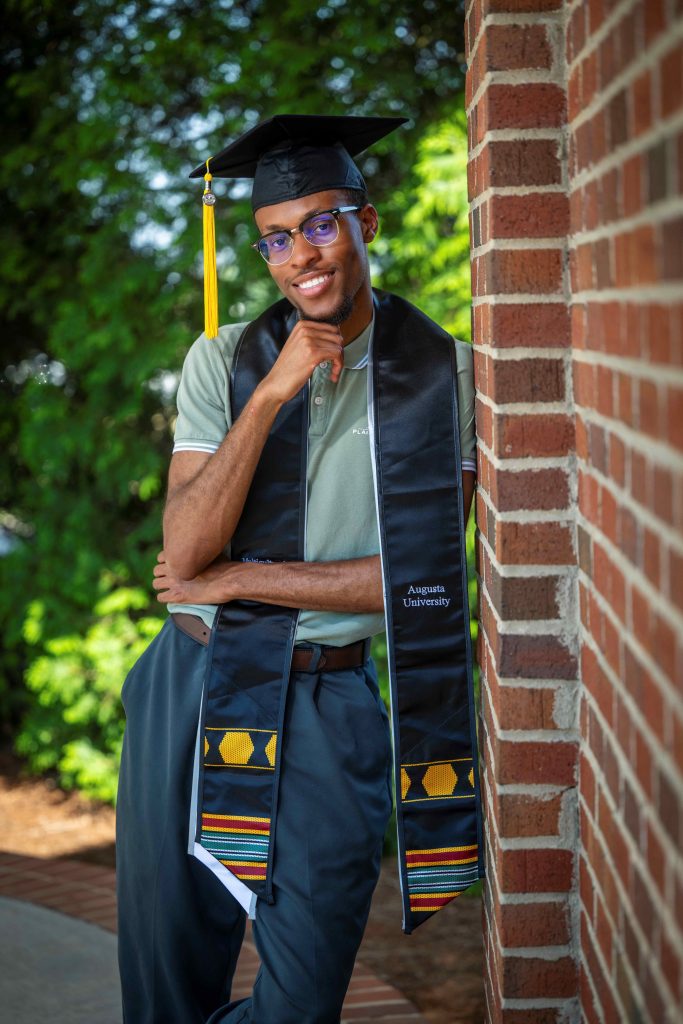 A man wearing a graduate hood and gown, while leaning against a brick wall, smiles for a photo. 