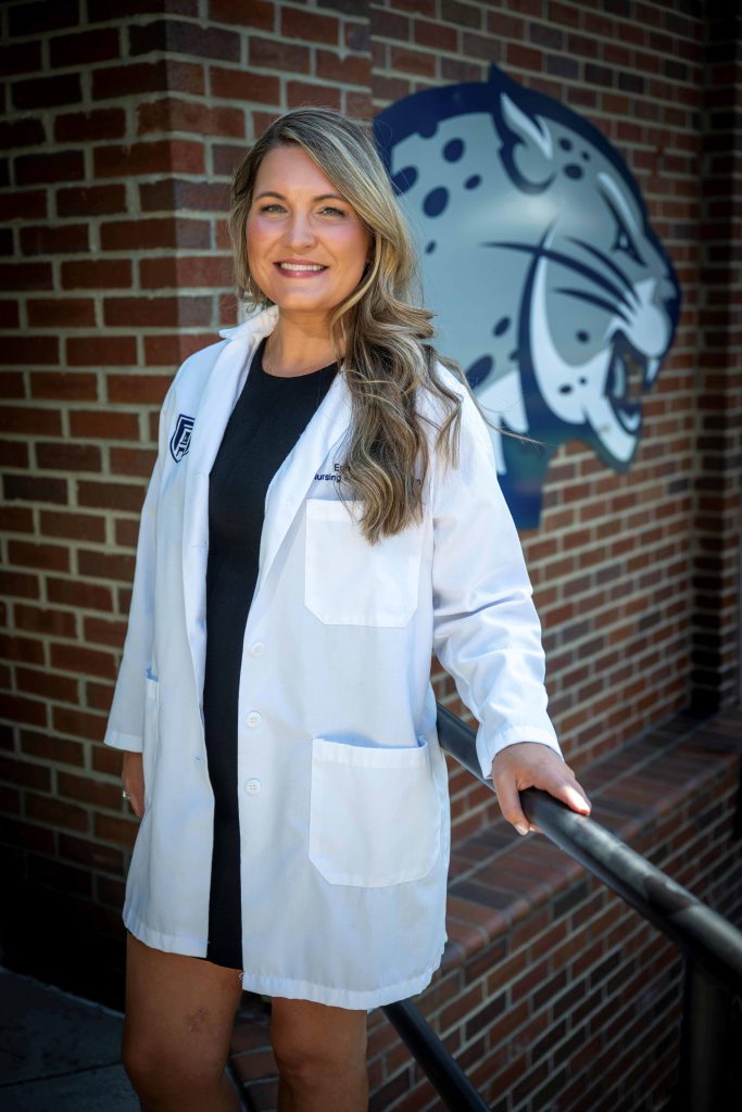 A woman in her white coat and a dress poses by a railing with a jaguar head logo behind her on a brick wall