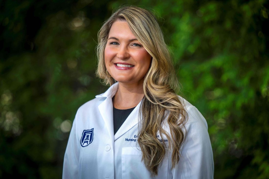 A woman in her lab coat smiles at the camera in front of greenery.