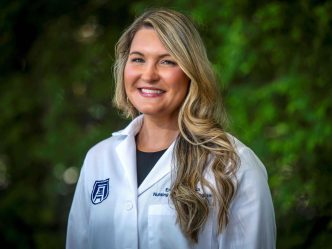 A woman in her lab coat smiles at the camera in front of greenery.