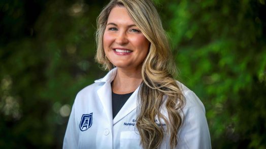 A woman in her lab coat smiles at the camera in front of greenery.