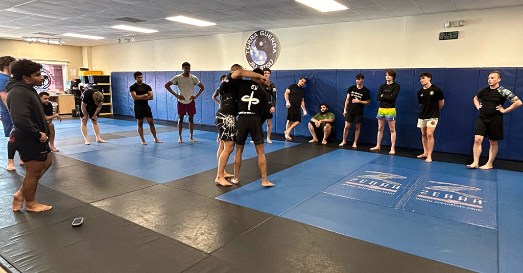 Two men Jiu-Jitsu spar on a mat while people observe.