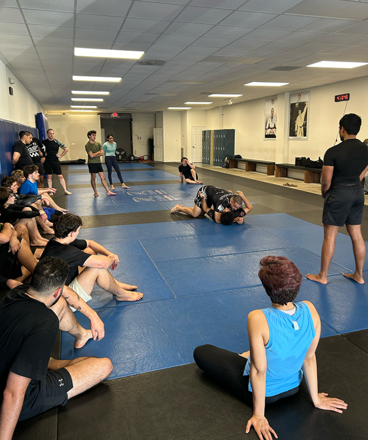 Two men Jiu-Jitsu spar on a mat while people observe.