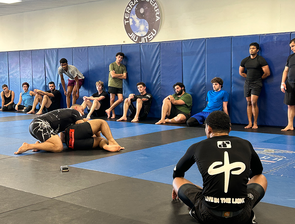 Two men demonstrate a Jiu-Jitsu technique on a mat while other men watch.