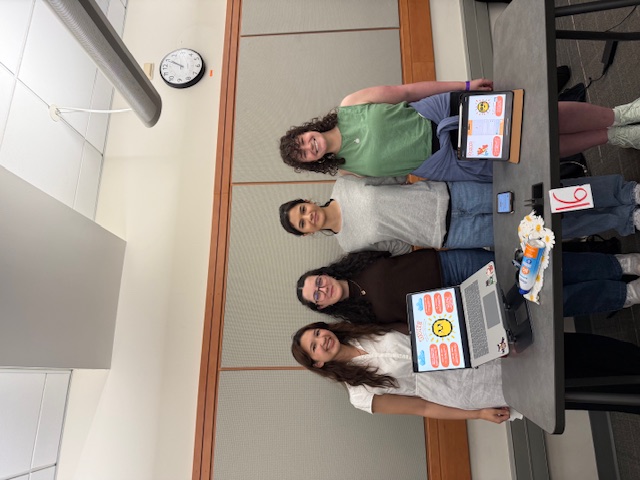 Four students stand behind a table displaying a project with a laptop, presentation board and materials in a classroom setting.