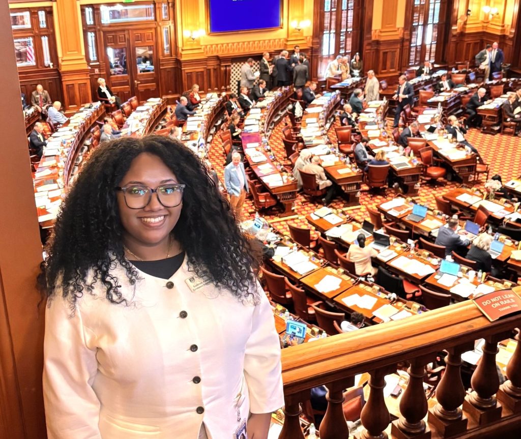 Standing on a stairwell above a floor chamber filled with desks, a young college-aged girl smiles for a photo.