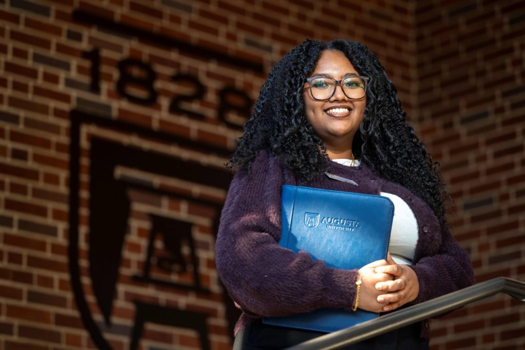 A young college-aged woman holding an Augusta University notebook smiles in front of a brick background. 