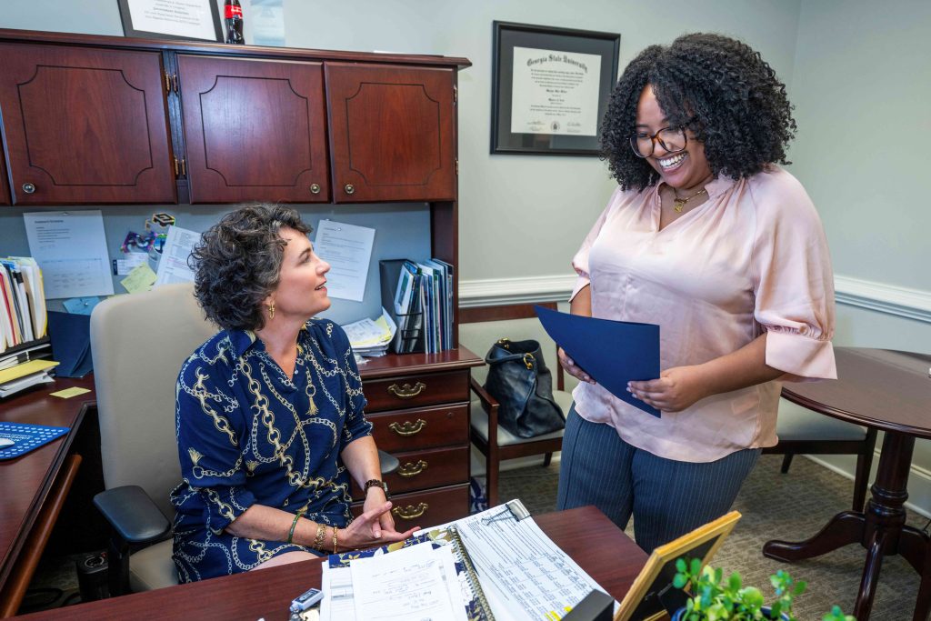 An older woman sitting at a desk smiling at a young college-aged woman holding a folder while standing in front of her.