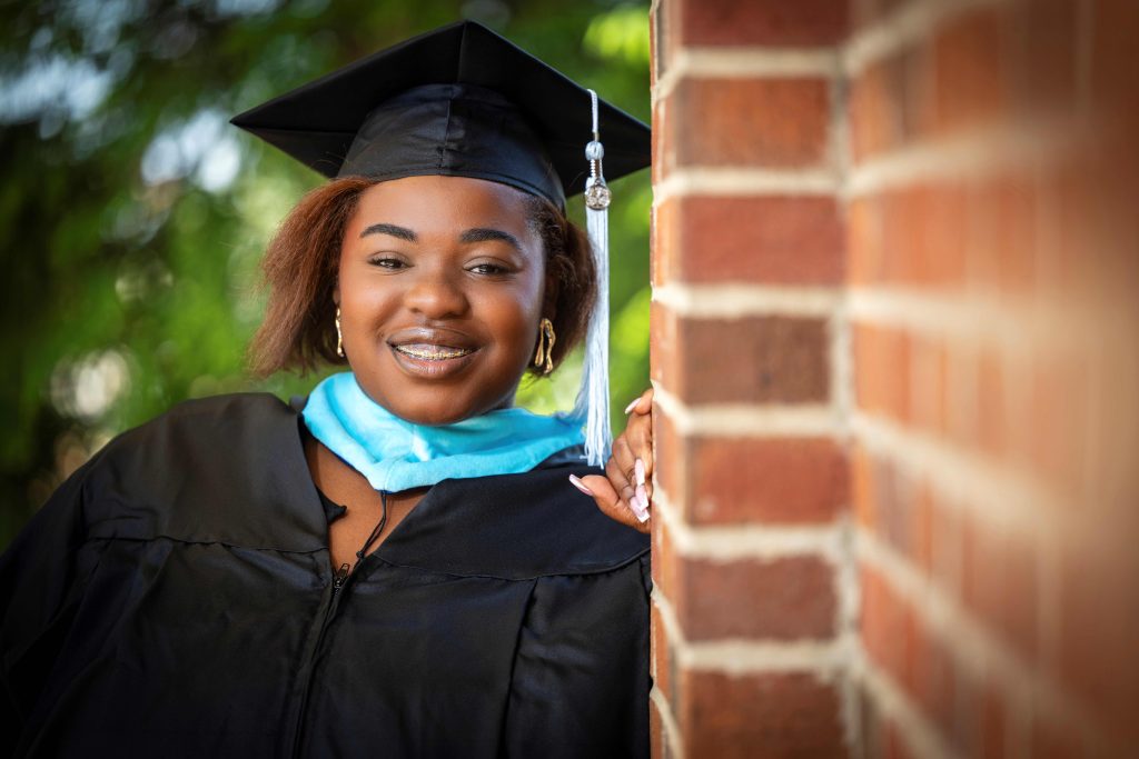 A woman wearing a graduate hood and gown, while leaning against a brick wall, smiles for a photo. 