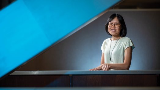 A female smiling and posing for a photo. Her hands are laying on a railing of some stairs.