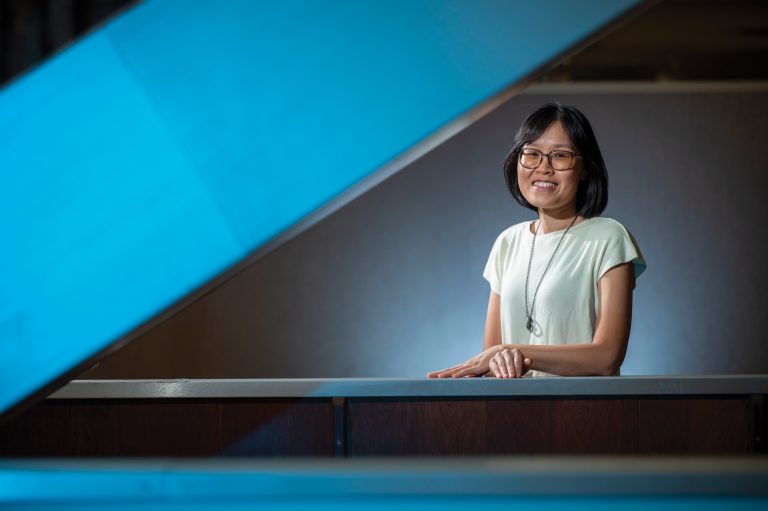 A female smiling and posing for a photo. Her hands are laying on a railing of some stairs.