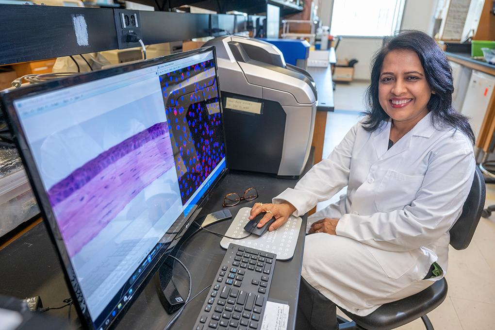 A female scientist wearing a lab coat sits at a computer and smiles at the camera.