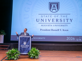 Man at podium using his hands in a speech with a large screen behind him.