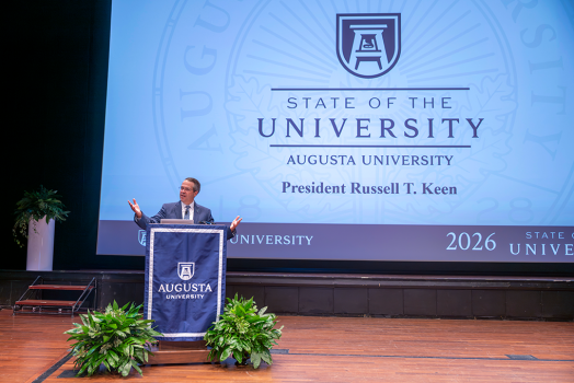 Man at podium using his hands in a speech with a large screen behind him.