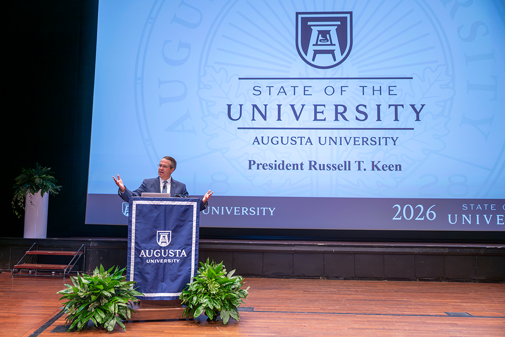 Man at podium using his hands in a speech with a large screen behind him.