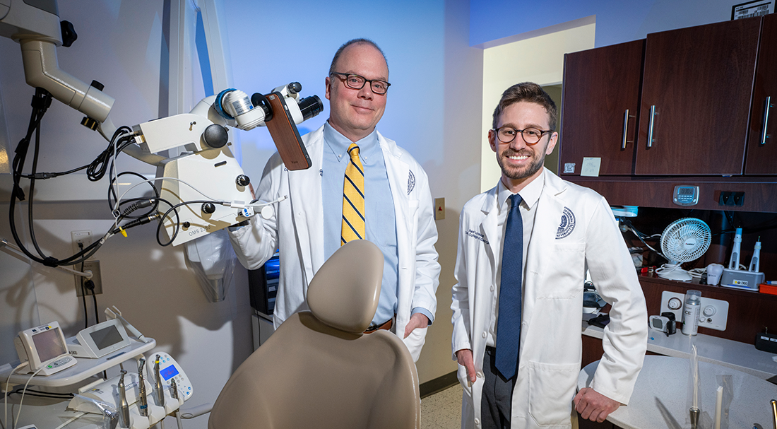 A dentist and a dental student wearing lab coats pose in an exam room surrounded by dental equipment.