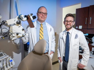 A dentist and a dental student wearing lab coats pose in an exam room surrounded by dental equipment.