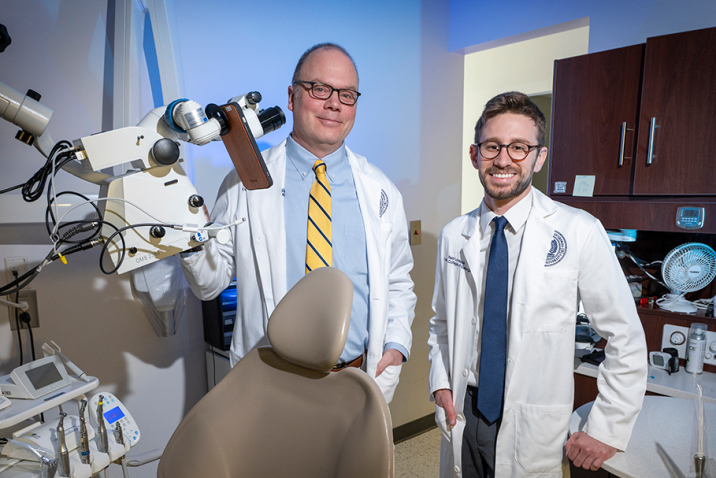 A dentist and a dental student wearing lab coats pose in an exam room surrounded by dental equipment.