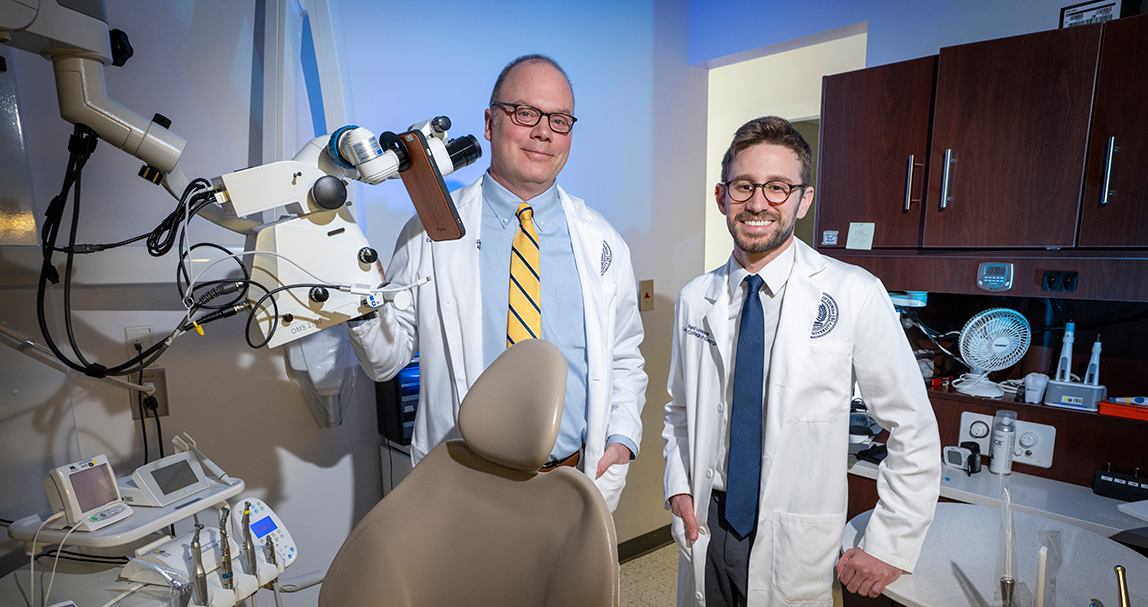 A dentist and a dental student wearing lab coats pose in an exam room surrounded by dental equipment.