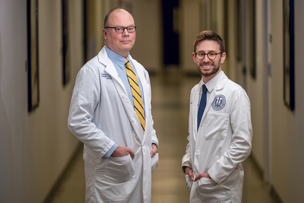 A dentist and a dental student wearing lab coats stand in a hallway and smile at the camera.