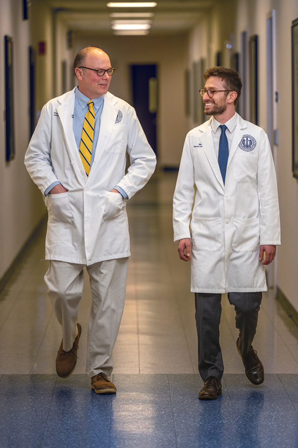 A dentist and a dental student wearing lab coats chat and stroll down a hallway.
