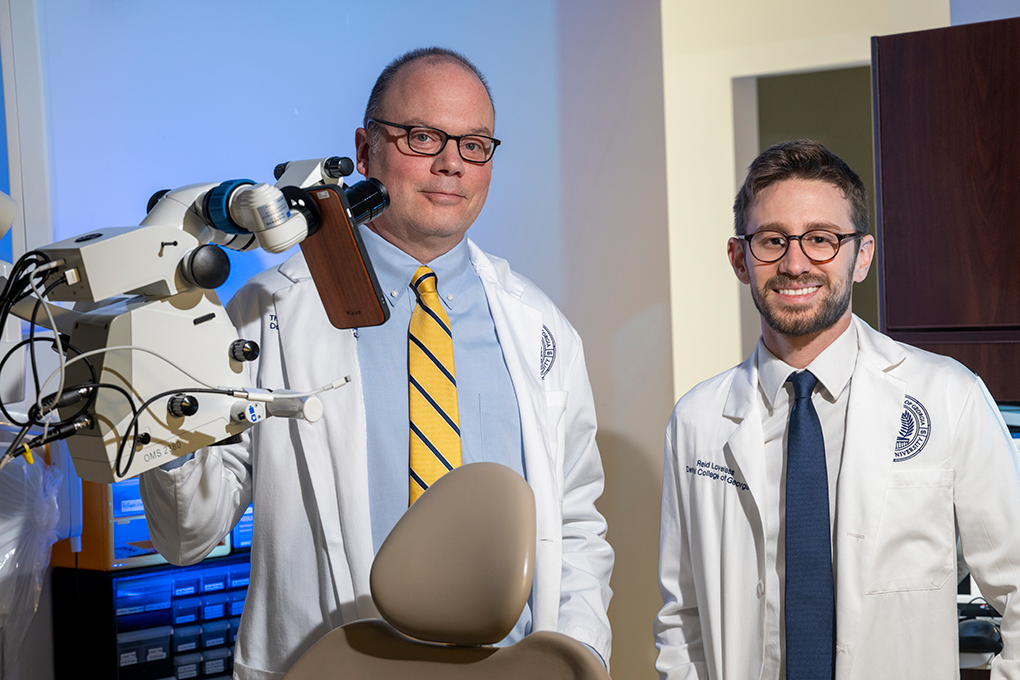A dentist and a dental student wearing lab coats pose for the camera in an exam room.