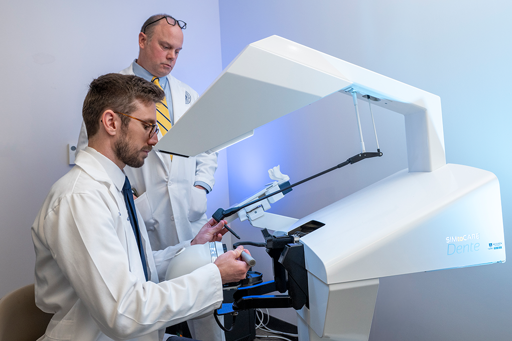 A dental student sits a dental treatment simulator while a supervising dentist observes him.