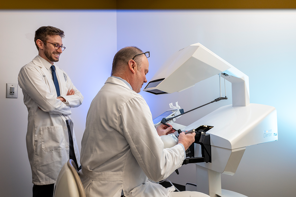 A dentist sits a dental treatment simulator while a dental student observes him.