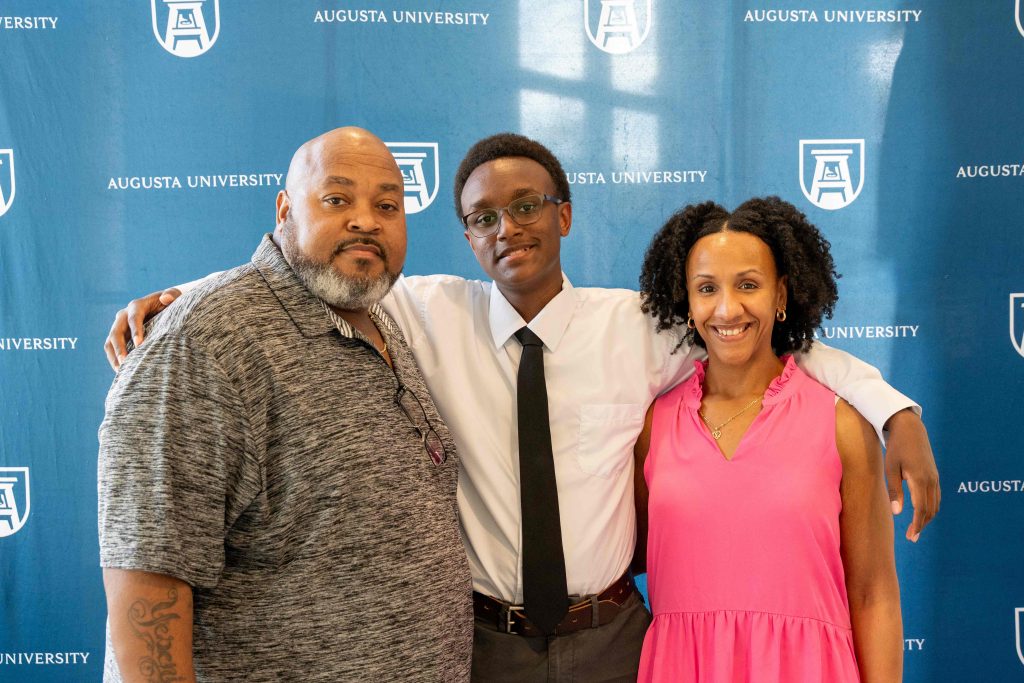 A high school-aged boy smiles for a photo, standing with his arms around an older man and woman.
