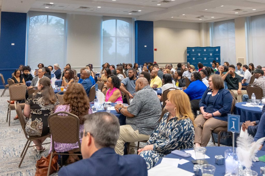 A large crowd of men and women sitting indoors at event tables.