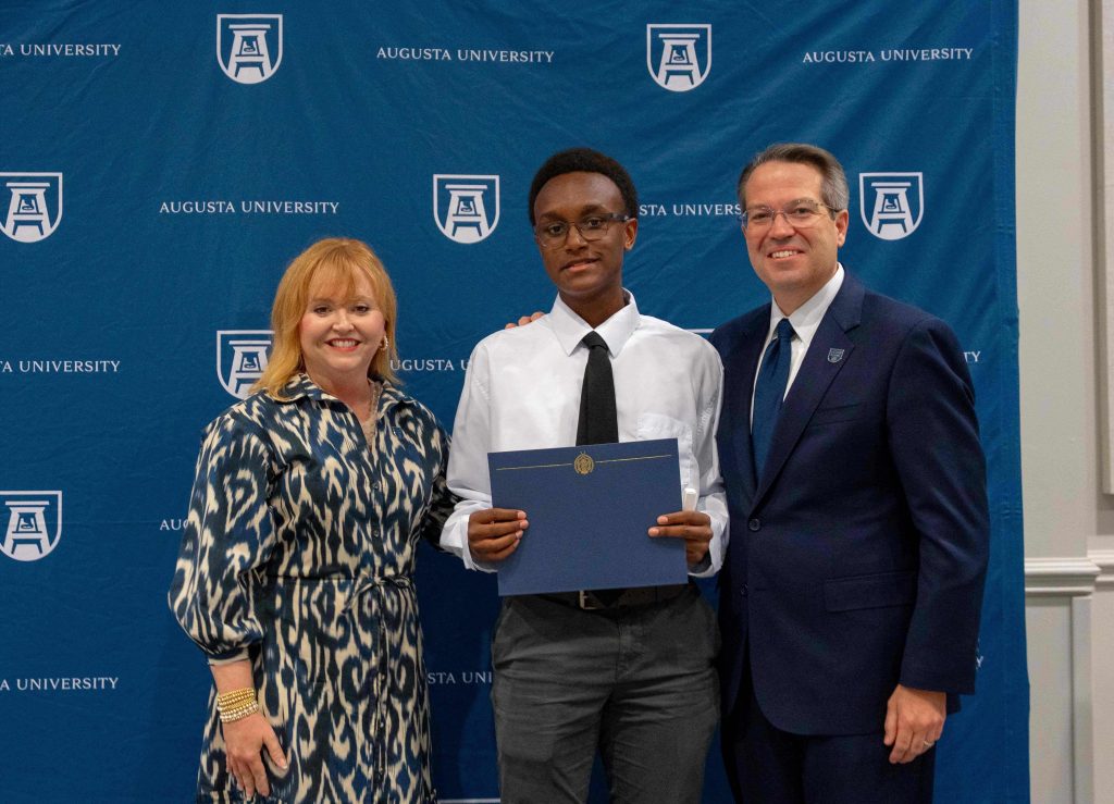 A high school-aged boy holds a certificate smiling between an older man and woman.