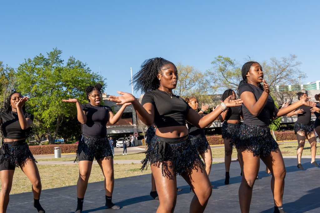 A group of girls in matching black outfits dance on an outdoor stage. 