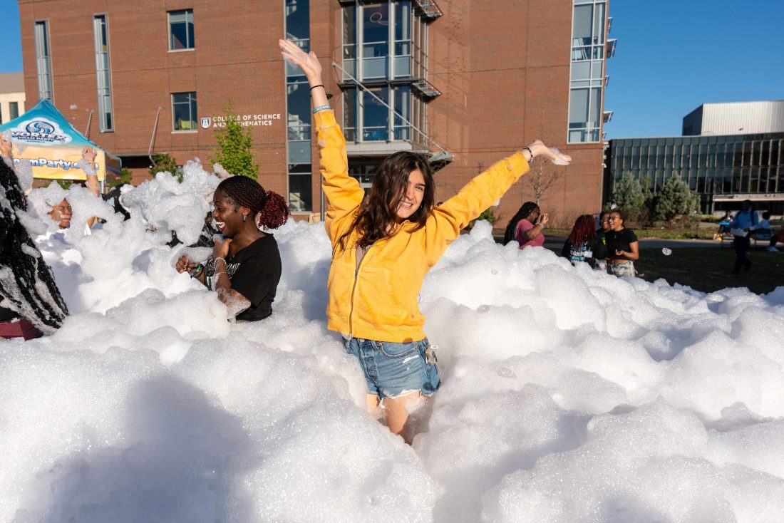 A girl poses with her arms up in the middle of a large amount of foam.