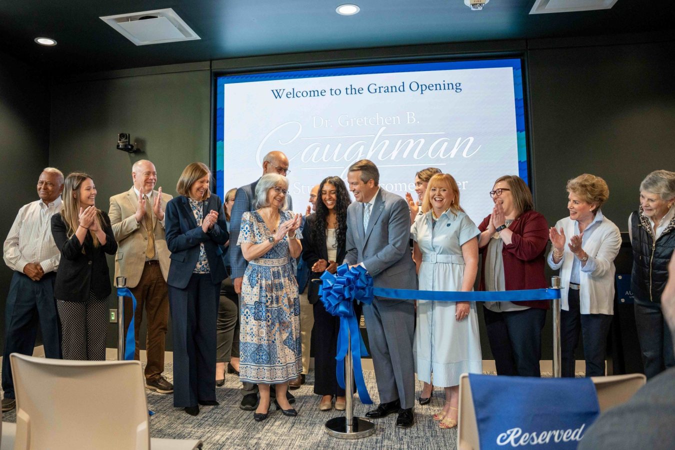 A large group of men and women gathered behind a ribbon at an indoor event.