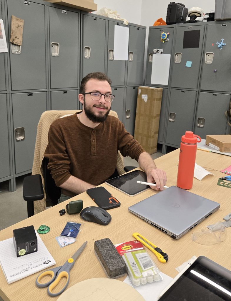 A young college-aged boy sitting at a desk with various items like laptop, water bottle and scissors on the table.