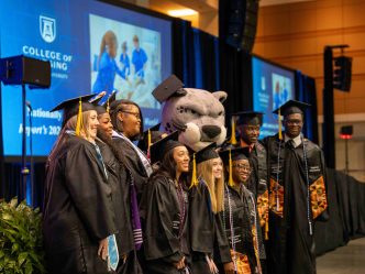 A large group of men and women wearing graduation caps and gowns pose for a photo with Augustus.