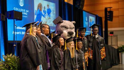 A large group of men and women wearing graduation caps and gowns pose for a photo with Augustus.