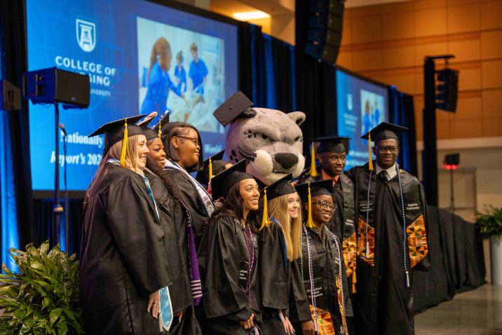 A large group of men and women wearing graduation caps and gowns pose for a photo with Augustus.