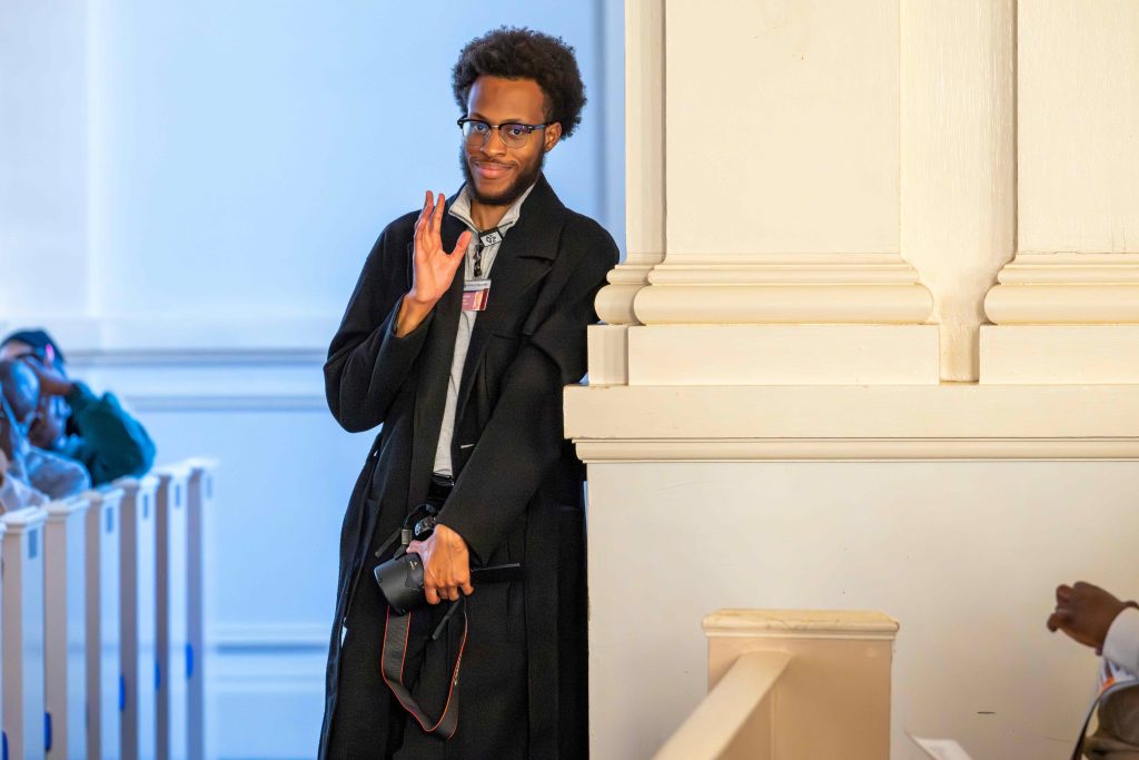 A college aged man holding a camera and waving at the photographer.