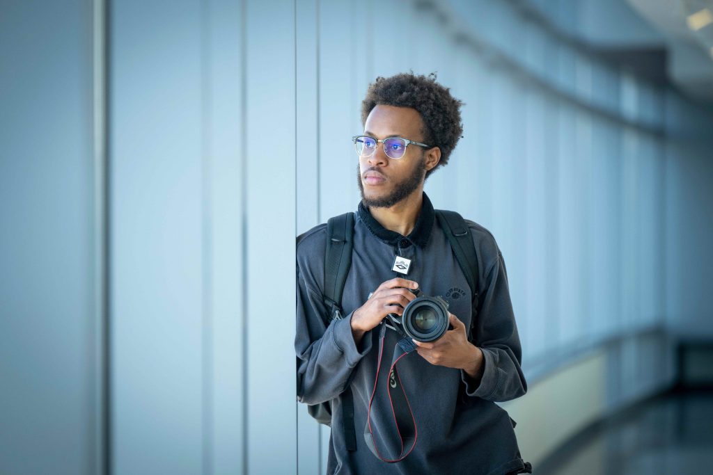 A college-aged man leans against a wall of windows while holding a camera.