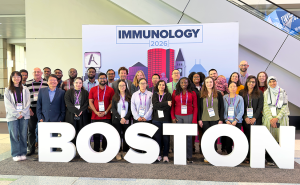A group of 28 people stand in a photo booth that includes oversized letters spelling out "Boston" in front of them and a backdrop that includes a silhouette of a cityscape and the words "Immunology 2026" behind them.