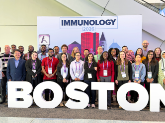 A group of 28 people stand in a photo booth that includes oversized letters spelling out "Boston" in front of them and a backdrop that includes a silhouette of a cityscape and the words "Immunology 2026" behind them.