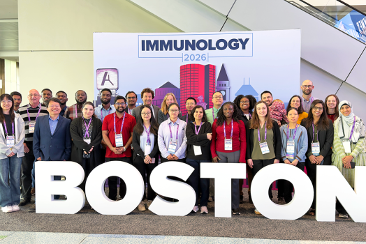 A group of 28 people stand in a photo booth that includes oversized letters spelling out "Boston" in front of them and a backdrop that includes a silhouette of a cityscape and the words "Immunology 2026" behind them.