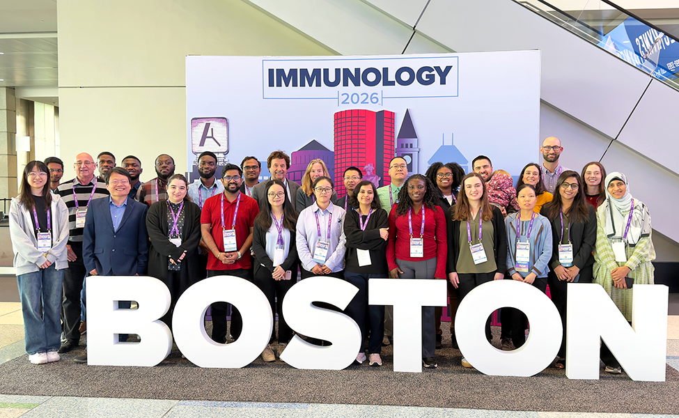 A group of 28 people stand in a photo booth that includes oversized letters spelling out "Boston" in front of them and a backdrop that includes a silhouette of a cityscape and the words "Immunology 2026" behind them.
