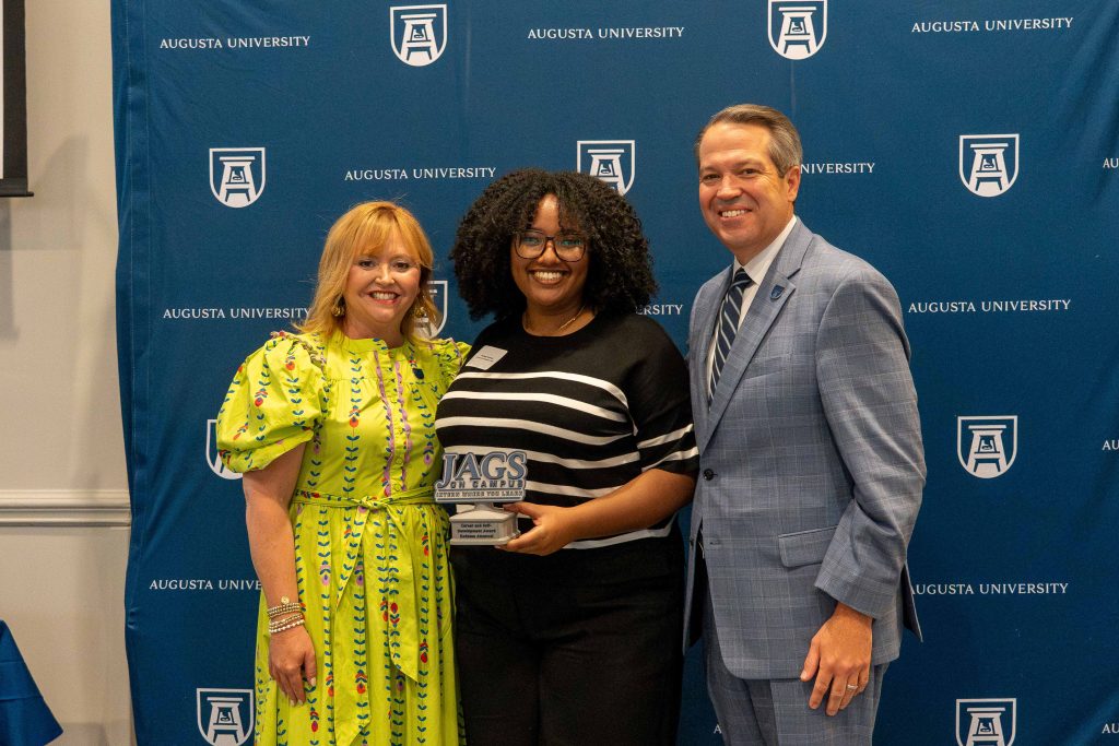 A college-aged girl holds an award, flanked by an older man on one side and an older woman on the other.