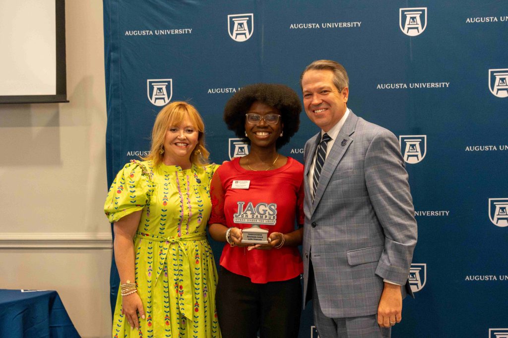 A college-aged girl holds an award, flanked by an older man on one side and an older woman on the other.